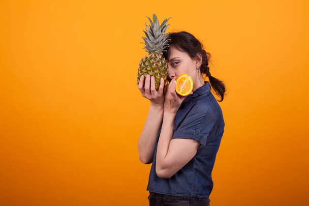 Visão lateral retrato de bela jovem segurando ananas saborosas e laranja em estúdio sobre fundo amarelo
 - Foto, Imagem