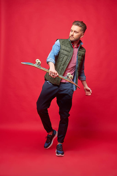 Fashionable man standing with skateboard in hand. Dynamic studio portrait, red background - Foto, Imagem