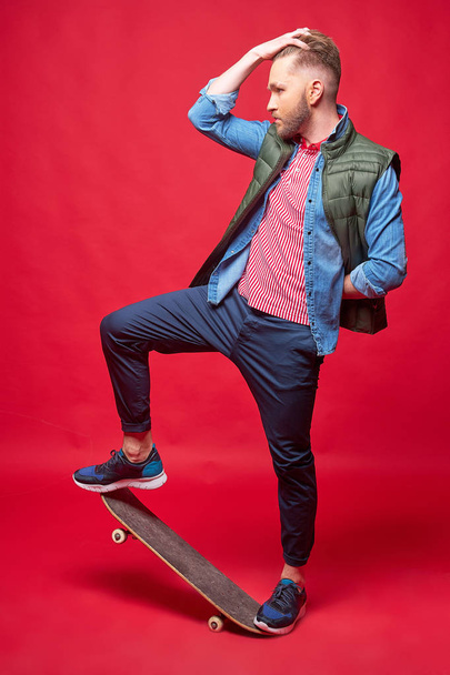 fashionable man in casual style standing on skateboard. Studio shot, red bacckground. - Photo, Image