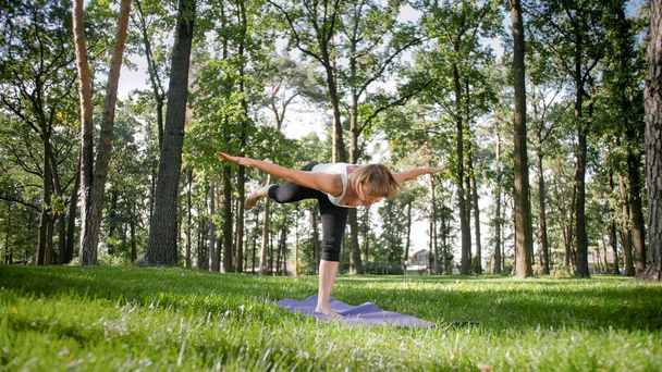 Zdjęcie w średnim wieku uśmiechnięta kobieta praktykujących jogę i medytowanie w parku. Kobieta rozciąganie i robi fitness na mat w lesie - Zdjęcie, obraz