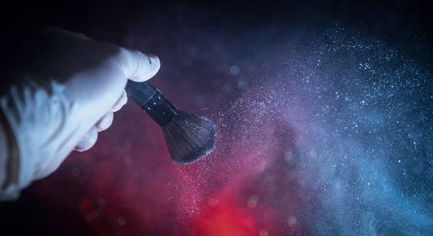 Makeup brush in hand with cosmetic powder on dark background with light and smoke. Powder splash on dark - Photo, Image
