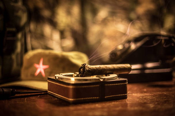 Közelről egy kubai szivart és hamutartó a fából készült asztal. Kommunista diktor parancsnok asztal sötét szobában. A hadsereg általános munkaasztal koncepciója. - Fotó, kép