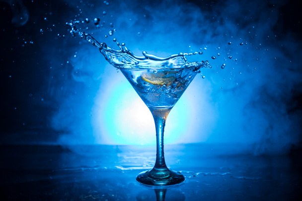 Martini-Cocktailglas in der Hand, das auf dunklem rauchigen Hintergrund plätschert, oder bunter Cocktail im Glas mit Spritzern und Oliven. Party Club Unterhaltung. gemischtes Licht. Selektiver Fokus - Foto, Bild