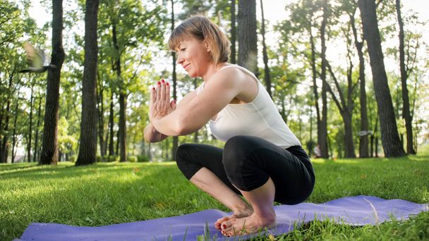 Image d'une femme d'âge moyen souriante en vêtements de fitness faisant des exercices d'étirement et de yoga. WOman méditer et faire du sport sur tapis de fitness sur herbe au parc
 - Photo, image