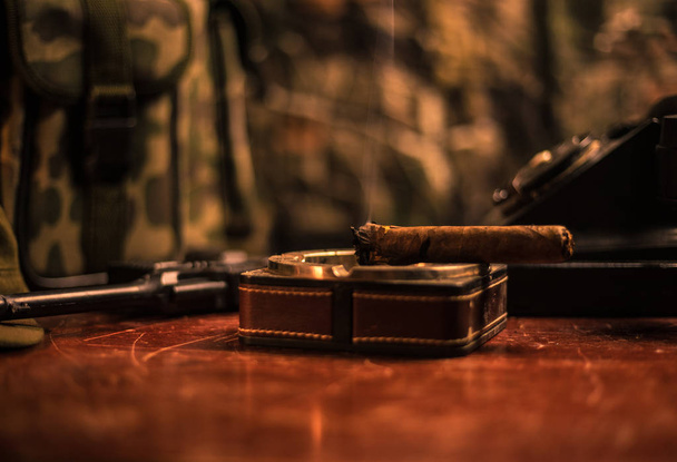 Nahaufnahme einer kubanischen Zigarre und eines Aschenbechers auf dem Holztisch. Kommandeurstisch des kommunistischen Diktators im dunklen Raum. Arbeitsplatzkonzept des Armeegenerals. - Foto, Bild