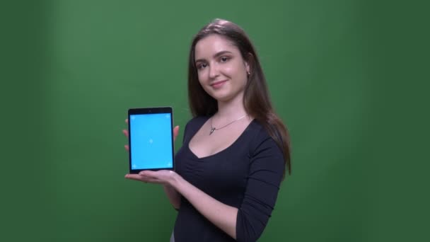 Closeup portrait of young pretty brunette female using the tablet and sshowing blue screen to camera with background isolated on green - Záběry, video