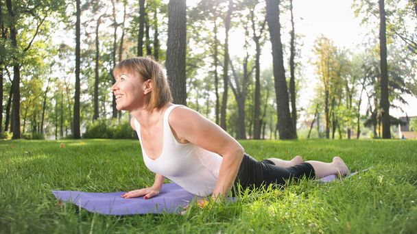 Obraz połowie wieku uśmiechnięty szczęśliwy kobieta medytuje i robi ćwiczenia jogi na trawie w lesie. Kobieta dbająca o jej zdrowie fizyczne i psychiczne podczas pracitising fitness i rozciągania w parku - Zdjęcie, obraz