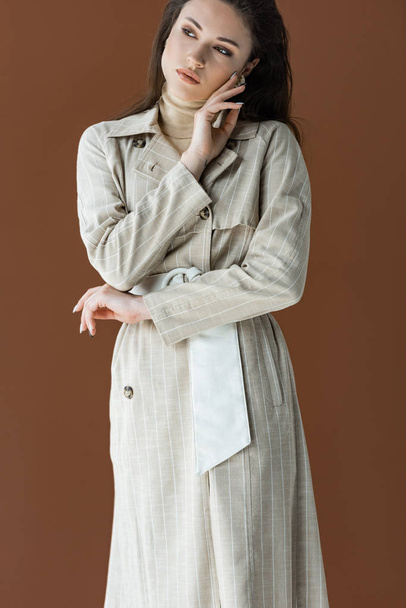 stylische Frau im trendigen Trenchcoat isoliert auf braun, die Hand vor dem Gesicht - Foto, Bild