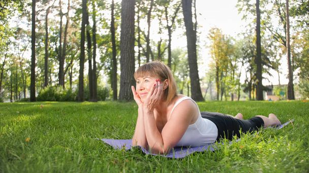 ストレッチやヨガのエクササイズをしているフィットネス服を着た中年女性の笑顔のイメージ。公園の芝生の上のフィットネスマットで瞑想し、スポーツをする女性 - 写真・画像