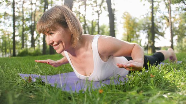 Photo of smiling happy woman 40 years old doing yoga exercises on fitness mat at forest. Harmony of human in nature. Middle aged people taking car of mental and physical health - Photo, Image