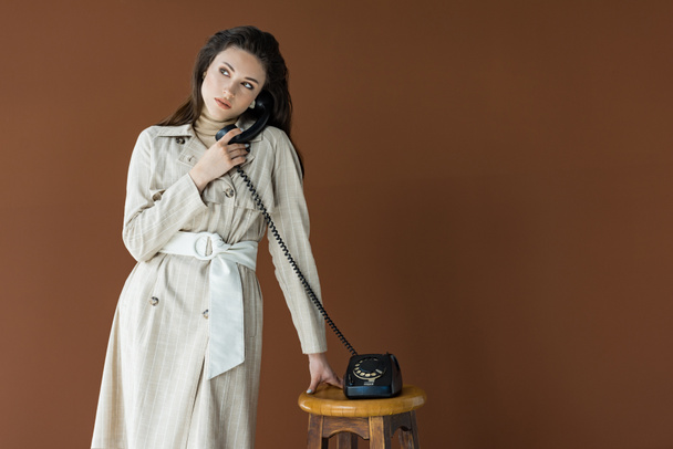 mujer elegante en gabardina hablando por teléfono, mirando hacia otro lado y de pie aislado en marrón
 - Foto, imagen