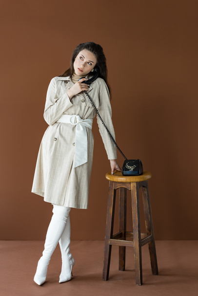 beautiful model in trench coat talking on telephone, looking away and standing isolated on brown - Photo, Image
