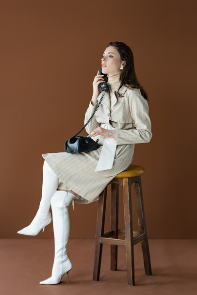 atraktivní model mluvení po telefonu, dívám se jinam a sedí izolovaně na hnědém - Fotografie, Obrázek