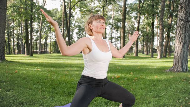 Obraz połowie wieku uśmiechnięty szczęśliwy kobieta medytuje i robi ćwiczenia jogi na trawie w lesie. Kobieta dbająca o jej zdrowie fizyczne i psychiczne podczas pracitising fitness i rozciągania w parku - Zdjęcie, obraz