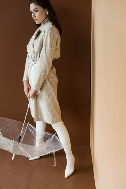 donna alla moda in elegante trench e stivali bianchi, che tiene l'ombrello, in piedi su sfondo marrone
 - Foto, immagini