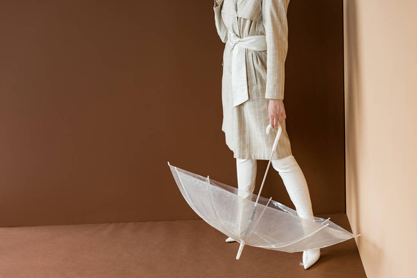 cropped view of model in beige trench holding umbrella, standing on brown background - Valokuva, kuva