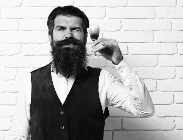 handsome bearded man with long beard and mustache has stylish hair on funny face holding glass of alcoholic shot in vintage suede leather waistcoat on white brick wall studio background - Fotoğraf, Görsel