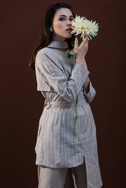 stylish model holding chrysanthemum flower near face, standing on brown background - Foto, afbeelding