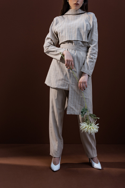 oříznutý pohled na módní model držící chryzantéum a stojící na hnědém pozadí - Fotografie, Obrázek