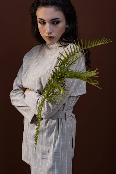 model holding fern leaves in hands, looking away, standing on brown background - Фото, зображення