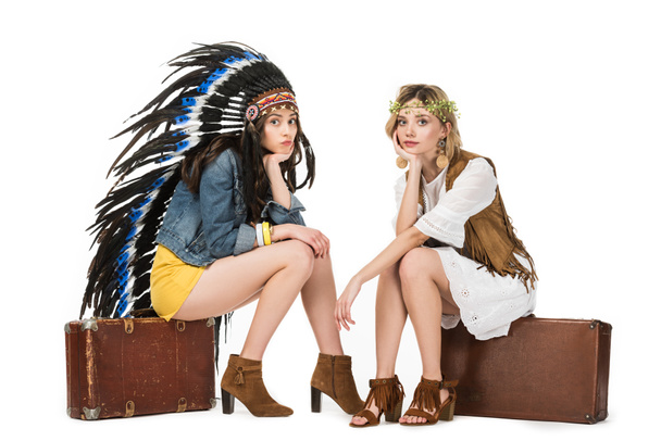 two bored bisexual hippie girls in indian headdress and wreath sitting on suitcases isolated on white - Photo, Image