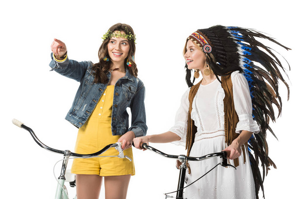 two smiling boho girls with bicycles pointing with finger isolated on white - Photo, Image
