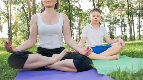 Photo of middle aged female yoga teacher or guru teaching teenage boy doing yoga. Woman with boy meditating and stretching on grass at park - Zdjęcie, obraz