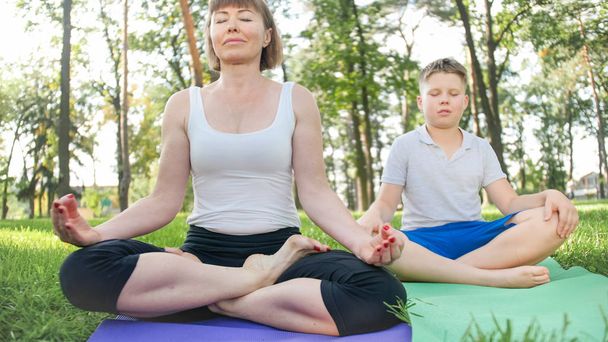 Portrait of 12 years old boy doing yoga exercise with his mother at park. Family meditating and stretching at forest - Photo, Image