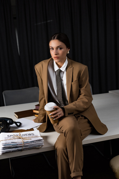 businesswoman in suit with paper cup sitting on table, crossing legs, looking at camera - Foto, imagen