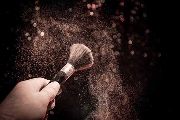 Make-up-Pinsel in der Hand mit Kosmetikpuder auf dunklem Hintergrund mit Licht und Rauch. Pulverspritzer auf Dunkelheit - Foto, Bild