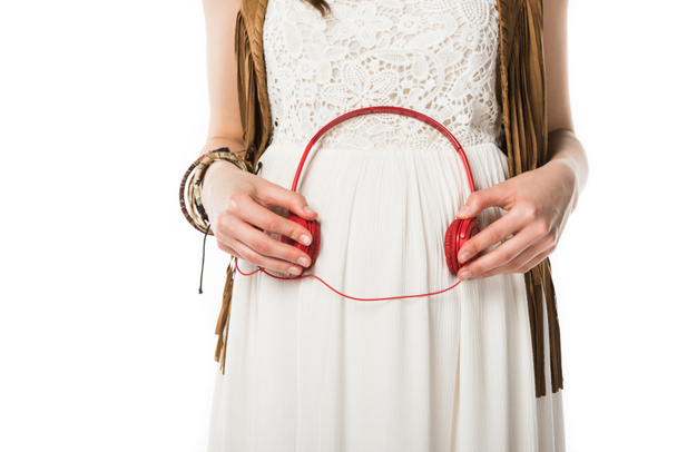 περικομμένη θέα της εγκύου χίπη γυναίκα κρατώντας ακουστικά στην κοιλιά απομονωθεί σε λευκό - Φωτογραφία, εικόνα