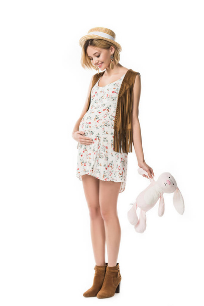 full length view of pregnant hippie girl in straw hat holding toy rabbit isolated on white - Valokuva, kuva