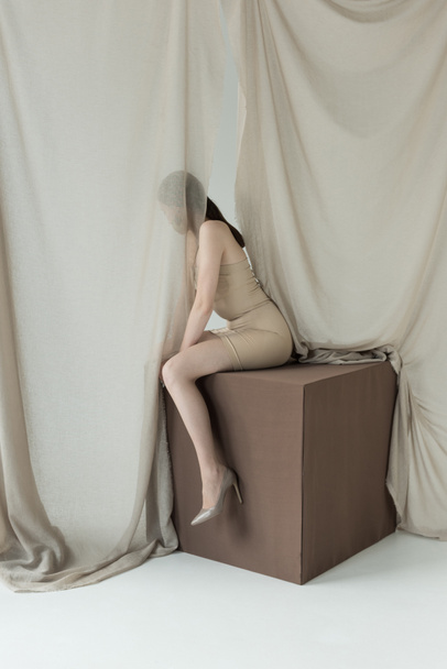 молодая модель в платье сидит на кубе за светлым занавесом
 - Фото, изображение