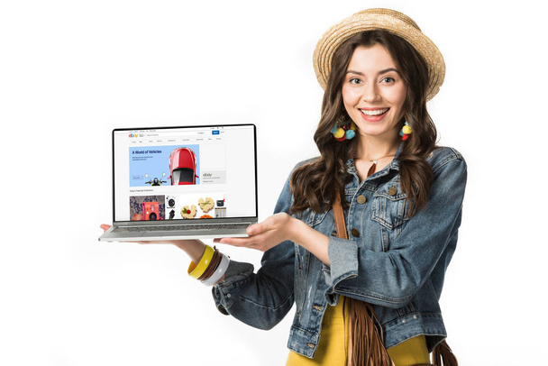 KYIV, UKRAINE - FEBRUARY 4, 2019: smiling hippie girl in straw hat holding laptop with ebay website on screen isolated on white - Foto, immagini