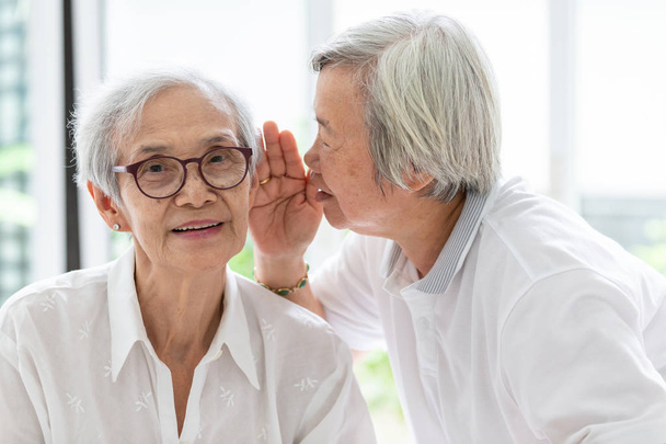 Asian senior woman holding hand near mouth telling funny,gossips on ears to friend,speaking in elderly woman ear and near face,having difficulty in hearing,hard to hear,hearing impairment,hearing impaired old people - Photo, Image