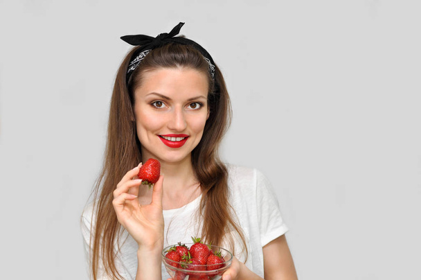 Beautiful sexy pin-up woman with bright makeup and red lipstick is eating red strawberries and smiling. - Photo, Image