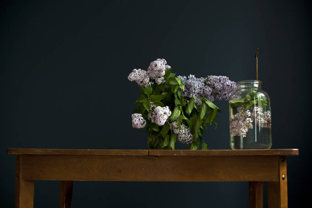 kytice fialových větviček v průhledné zelené nádobě na dřevěném stole naproti černé stěně - Fotografie, Obrázek