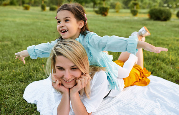 Happy child playing with pretty mother in the park. Beautiful young woman and her daughter relaxing on the green grass. Mom and little girl shares love. Happy Mothers Day. Motherhood and childhood - Foto, afbeelding