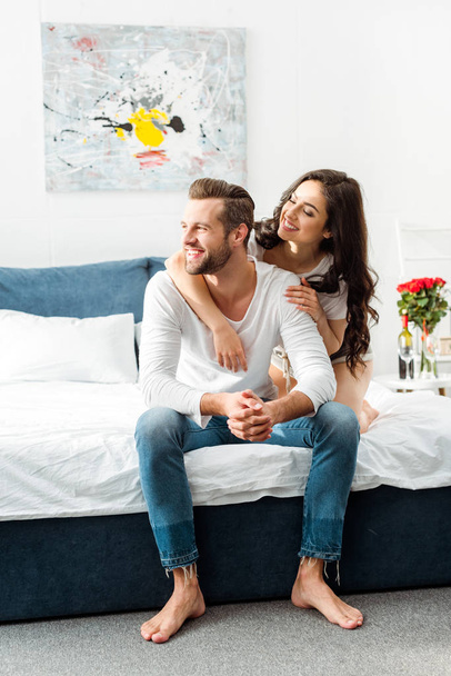 smiling girlfriend hugging boyfriend in bed at morning while man looking away - Photo, Image