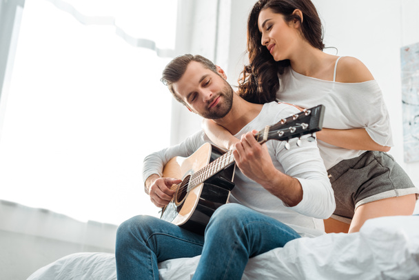 pohled nízkého úhlu pohledu na člověka hrající na akustickou kytaru poblíž atraktivní ženy v posteli - Fotografie, Obrázek