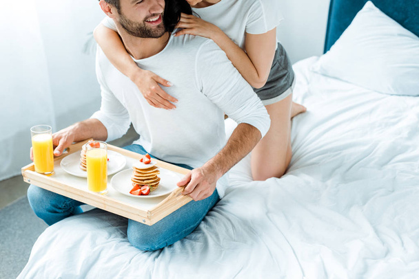 ベッドの上のトレイにオレンジジュースとパンケーキを持つ男を抱きしめる女性のトリミングされたビュー - 写真・画像
