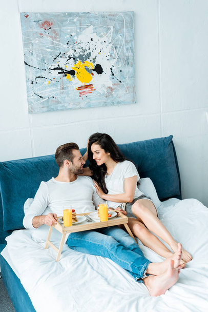 lächelndes glückliches Paar mit Orangensaft und Pfannkuchen zum Frühstück im Schlafzimmer - Foto, Bild