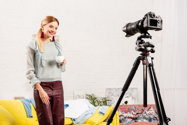 smiling blogger in earrings holding cup of coffee in front of video camera at home - Foto, Bild