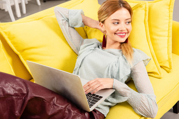 黄色いソファに横たわり、自宅でラップトップを使用して笑顔のフリーランサー - 写真・画像