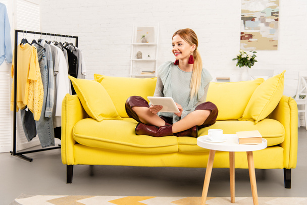 lächelndes Mädchen auf gelbem Sofa mit gekreuzten Beinen und mit digitalem Tablet - Foto, Bild