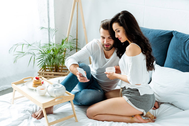 happy couple having breakfast in bed at morning and using smartphone - Photo, Image