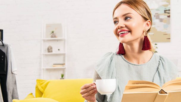 smiling girl in earrings holding cup of coffee and book at home - Photo, Image