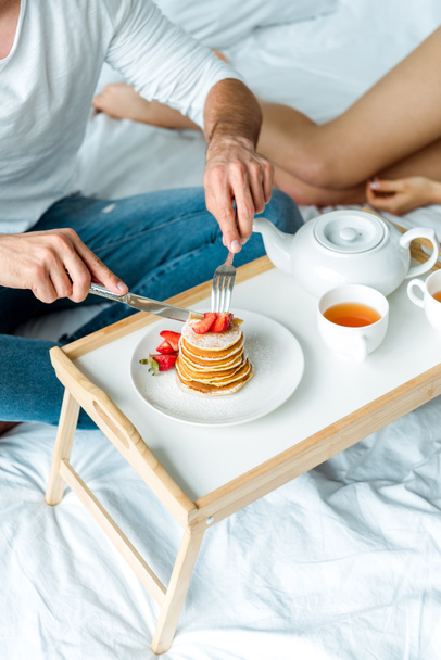 abgeschnittene Ansicht eines Mannes, der Pfannkuchen mit Erdbeeren isst und Tee im Bett trinkt - Foto, Bild