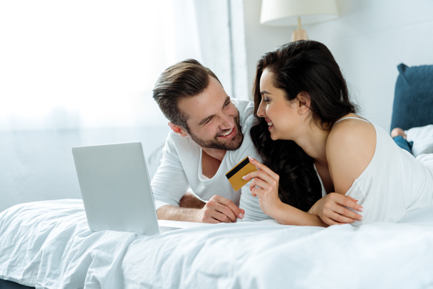 happy couple lying in bed near laptop while woman holding credit card, illustrative editorial - Foto, Imagem