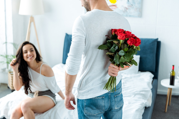 selective focus of happy girlfriend looking at boyfriend with red roses behind back  - Fotoğraf, Görsel